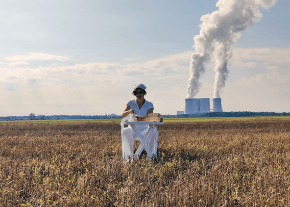 Foto: Eine Frau in weiß, sitzt auf einen abgeernteten Feld vor einem Tisch, im Hintergrund ein großes Kohlekraftwerk