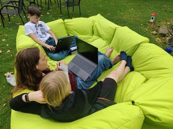 Drei Jugendliche mit Laptops, die gemütlich auf einem großen grünen Sitzsacksofa auf der Wiese herumlümmeln