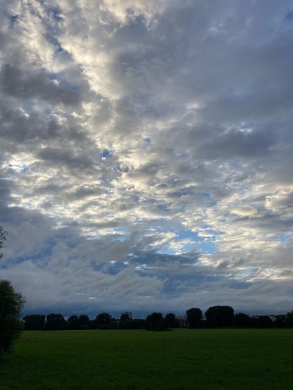 Wolken werden von der Sonne von hinten angeleuchtet. Weiße flockige Wolken vor blauem Himmel und wegziehenden Regenwolken. Im Vordergrund eine Wiese mit einer weiter hinten stehenden Baumreihe.
