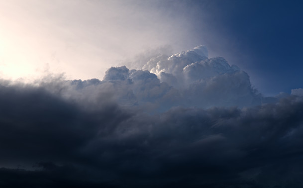 A dramatic and breathtaking cloudscape during the last moments of daylight. The image showcases a stunning contrast between the bright, illuminated upper clouds and the dark, looming lower clouds of a thrunderstorm. The upper part of the sky is bathed in a soft, ethereal light, creating a beautiful gradient that transitions from a bright, almost white hue to a deeper blue. The upper clouds are thick and billowy, with their edges glowing softly from the light behind them. In contrast, the lower clouds are darker and more ominous, creating a sense of depth and drama in the scene. The overall effect is a striking visual representation of the interplay between light and shadow, highlighting the beauty and power of nature.