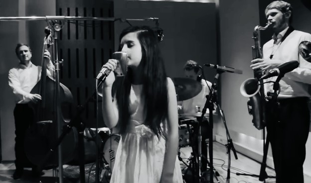 Screenshot aus dem Video in schwarz-weiß. In der Mitte die junge Angelina im Studio. Im Hintergrund drei Musiker.