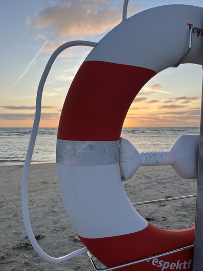 Ein Sonnenuntergang in hellen Rot-Orange-Tönen, etwas bewegte Nordsee, ein Stück Strand, durch einen Rettungsring fotografiert (im Vordergrund)