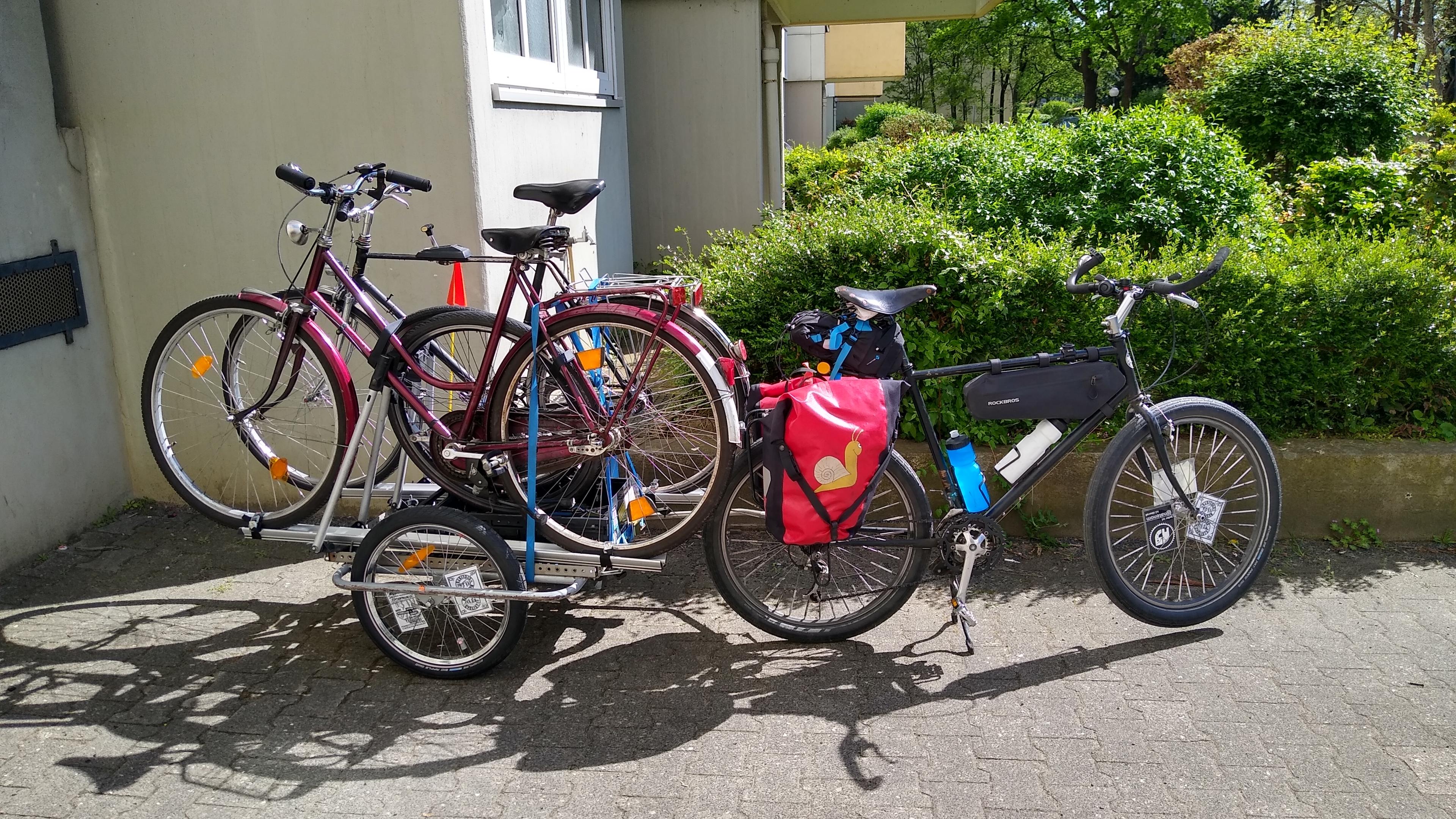 Ein schwarzes Fahrrad mit roten Packtaschen, daran ein Anhänger auf den mit fürs Auto gedachten Dachträgern zwei Fahrräder aufgeladen sind. Ein schwarzes Fahrrad mit roten Packtaschen, daran ein Anhänger auf den mit fürs Auto gedachten Dachträgern zwei Fahrräder aufgeladen sind.