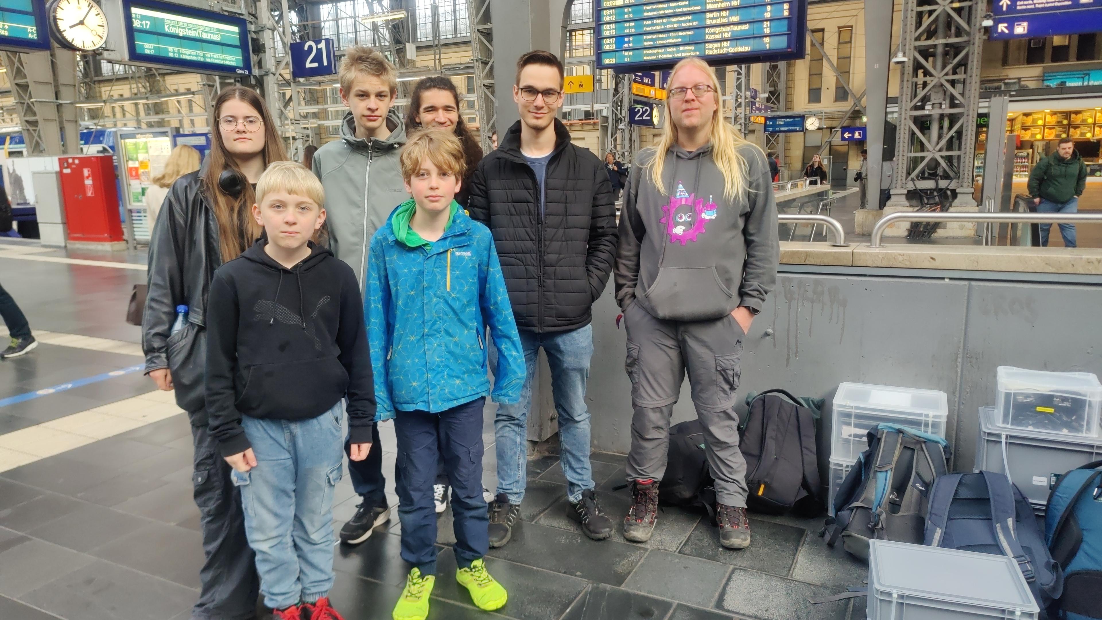Mehrere Kinder, Jugendliche und Erwachsene stehen mit Euroboxen und Rucksäcken an einer Treppe am Bahnhof.