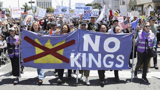 Bild eines Demonstrationszuges von vorne, bei dem sieben bis acht  Menschen  ein großes Banner mit der Aufschrift "No Kings" und einer durchgekreuzten Krone tragen