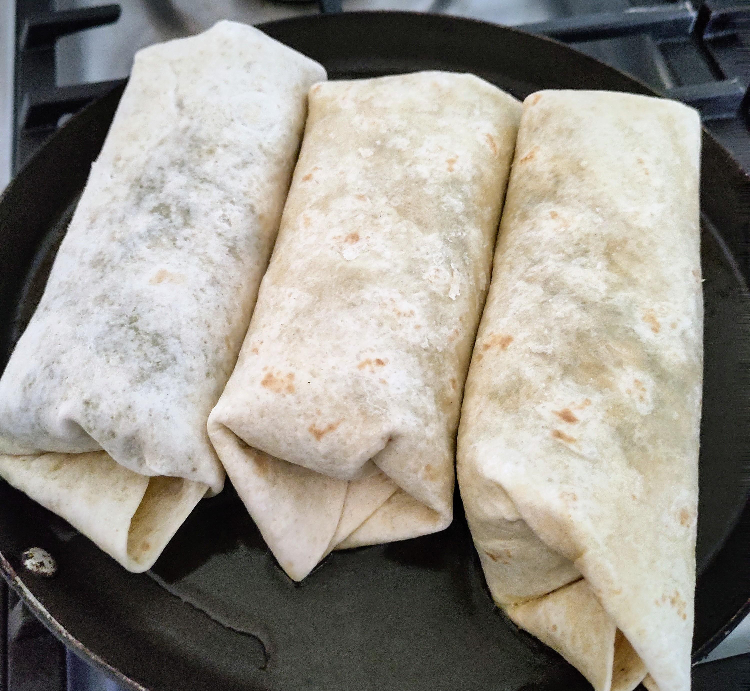 Drei ungebräunte, gewickelte Burritos liegen nebeneinander in einer Pfanne auf einem Herd. Die Burritos bestehen aus hellem Weizentortilla und sind bereit zum 
Braten.
