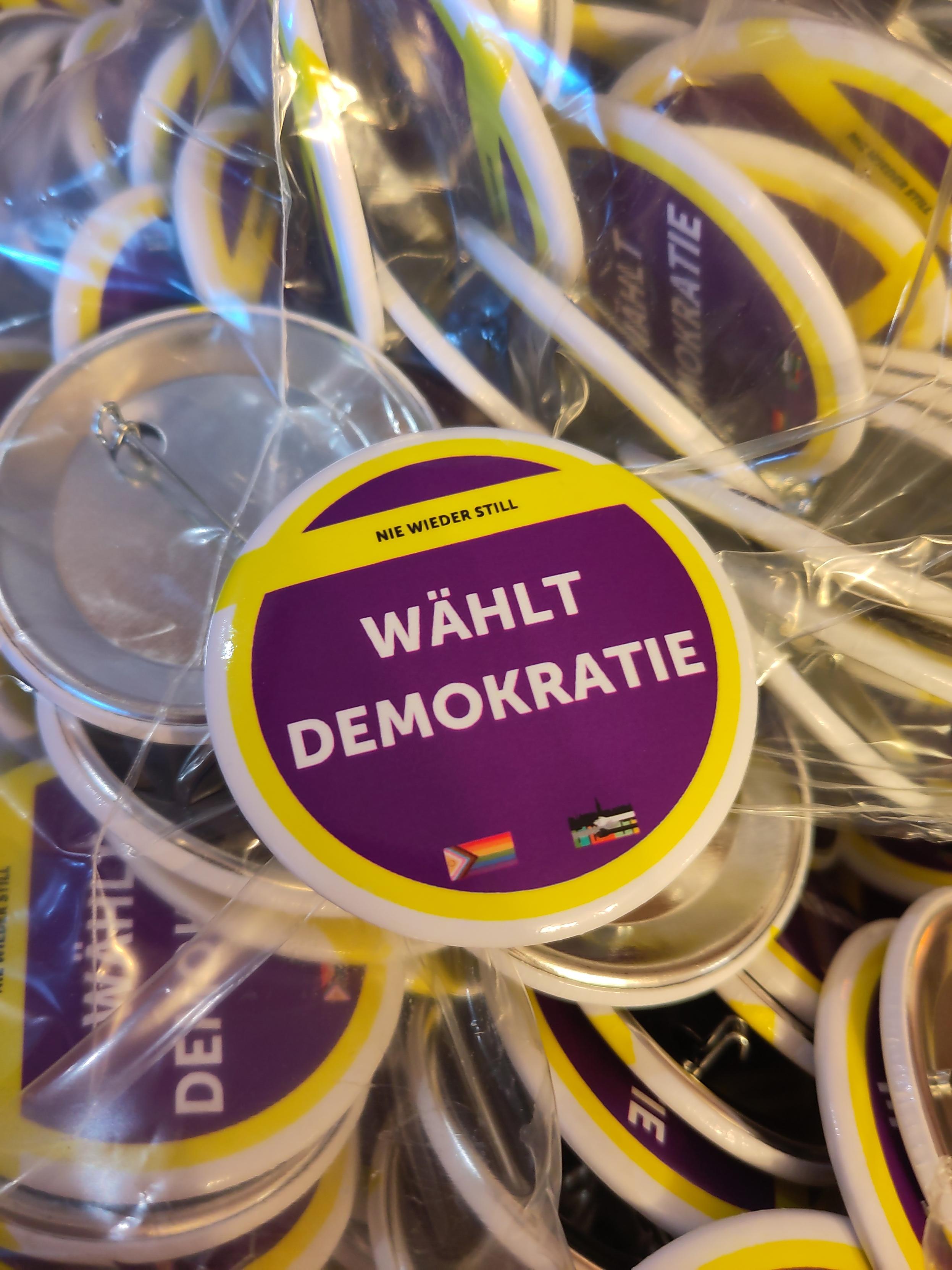 Button mit Aufschrift "Wählt Demokratie" in gelb, lila. Kleine Progressflagge und Logo des Schorndorfer Bündnissses gegen Rassismus und Rechtsextremismus