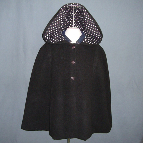 Cape à capuche en velours de laine noir doublée de coton à pois blancs sur fond noir fermée par pressions.