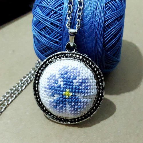 Un collier chaine à maille avec un grand pendentif en métal argenté broderies sur toile aida fleur bleu brodé